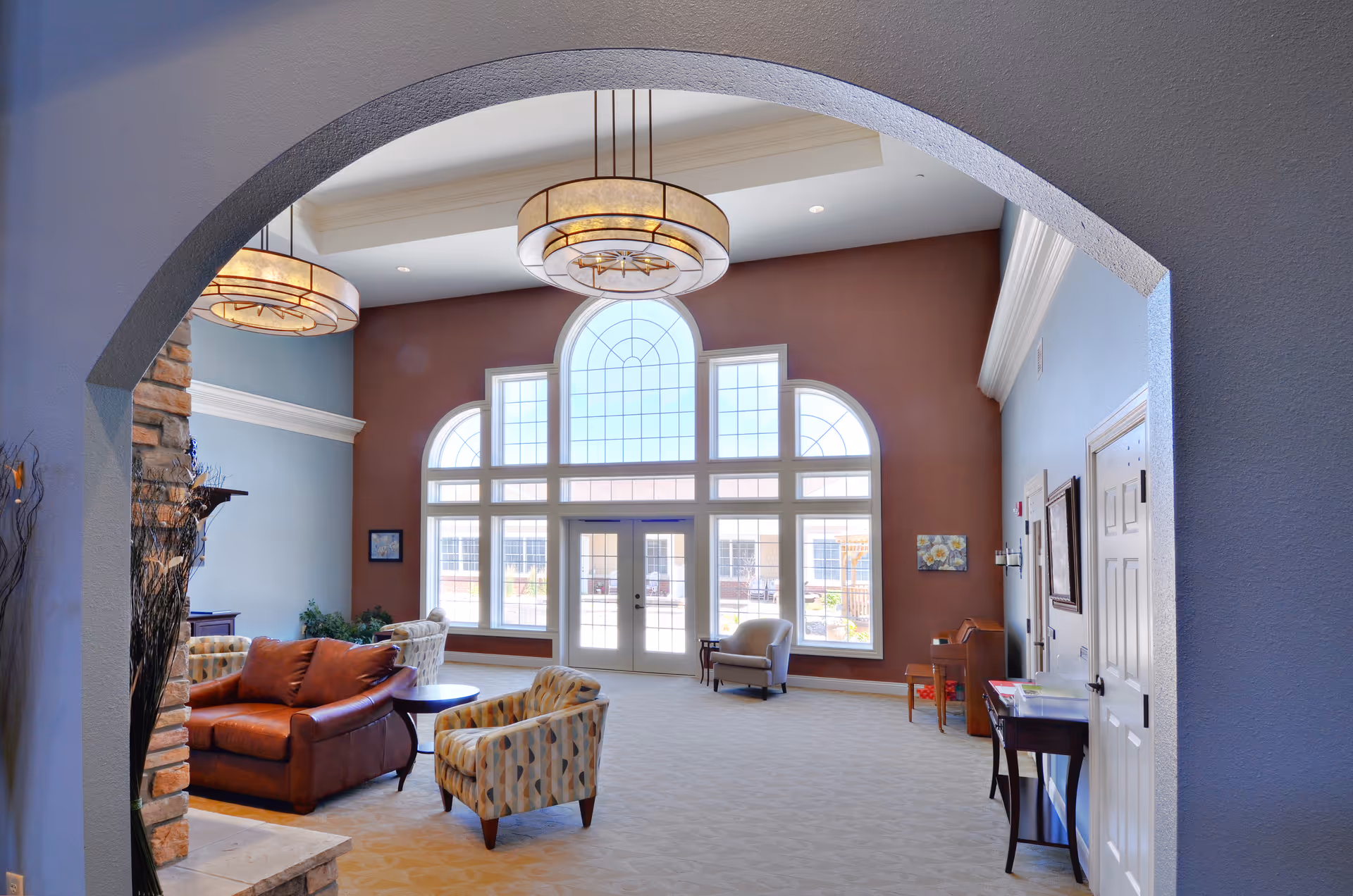 Spacious and well-lit living room area in Snyder Village Assisted Living with large arched windows, high ceilings, two modern hanging light fixtures, a brown leather sofa, patterned armchairs, and a stone fireplace on the left side.