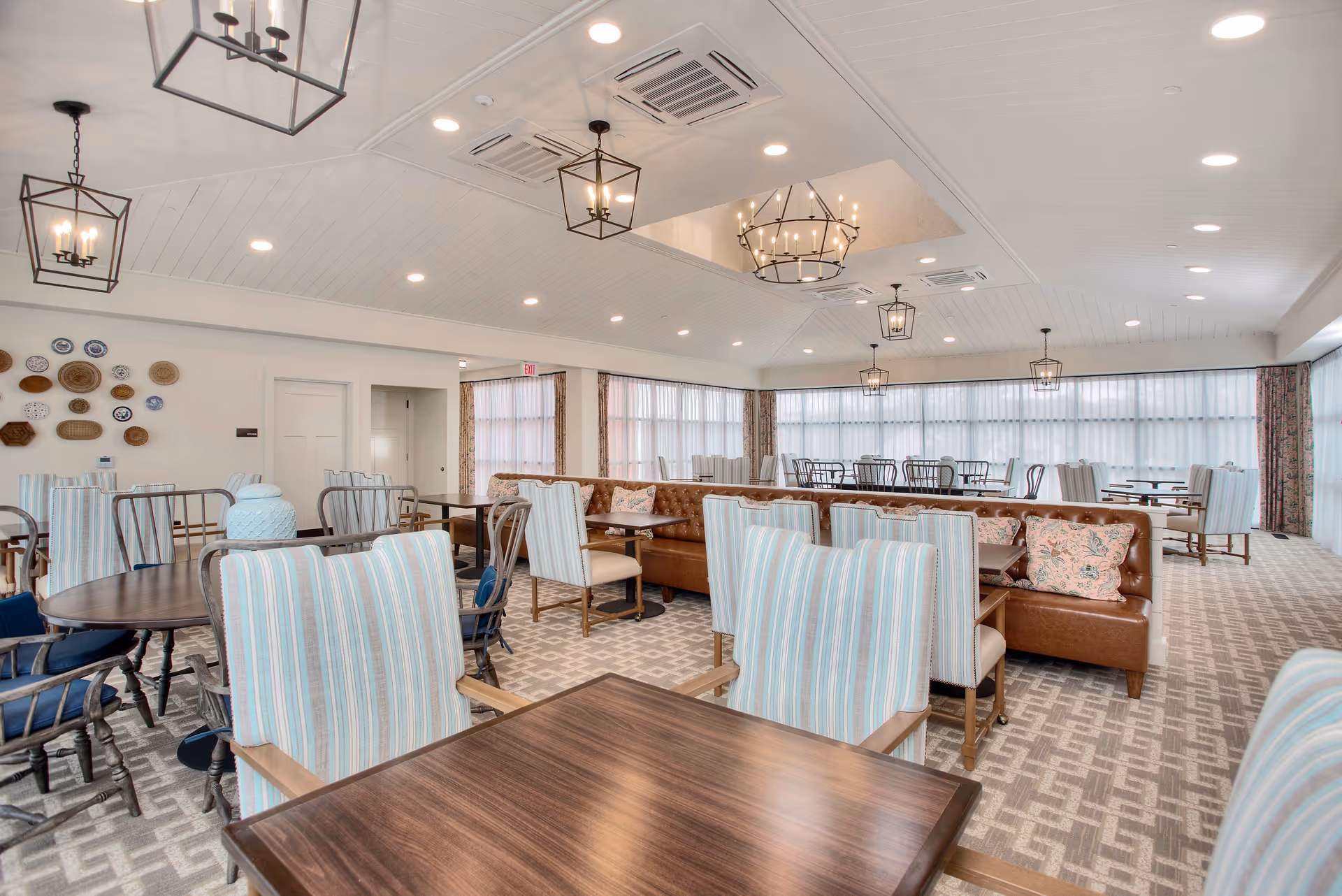 A spacious dining room with multiple tables and chairs, featuring striped upholstered chairs and a long brown leather bench with floral pillows. The room has large windows with sheer curtains, patterned carpet, and several modern and rustic light fixtures hanging from a white paneled ceiling.