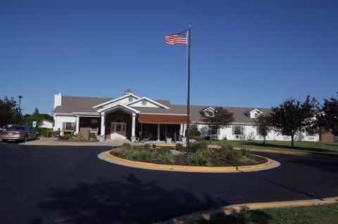 Front exterior view of Oakley Courts Assisted Living Community building with a circular driveway, landscaped garden in the center, an American flag on a flagpole, and clear blue sky.