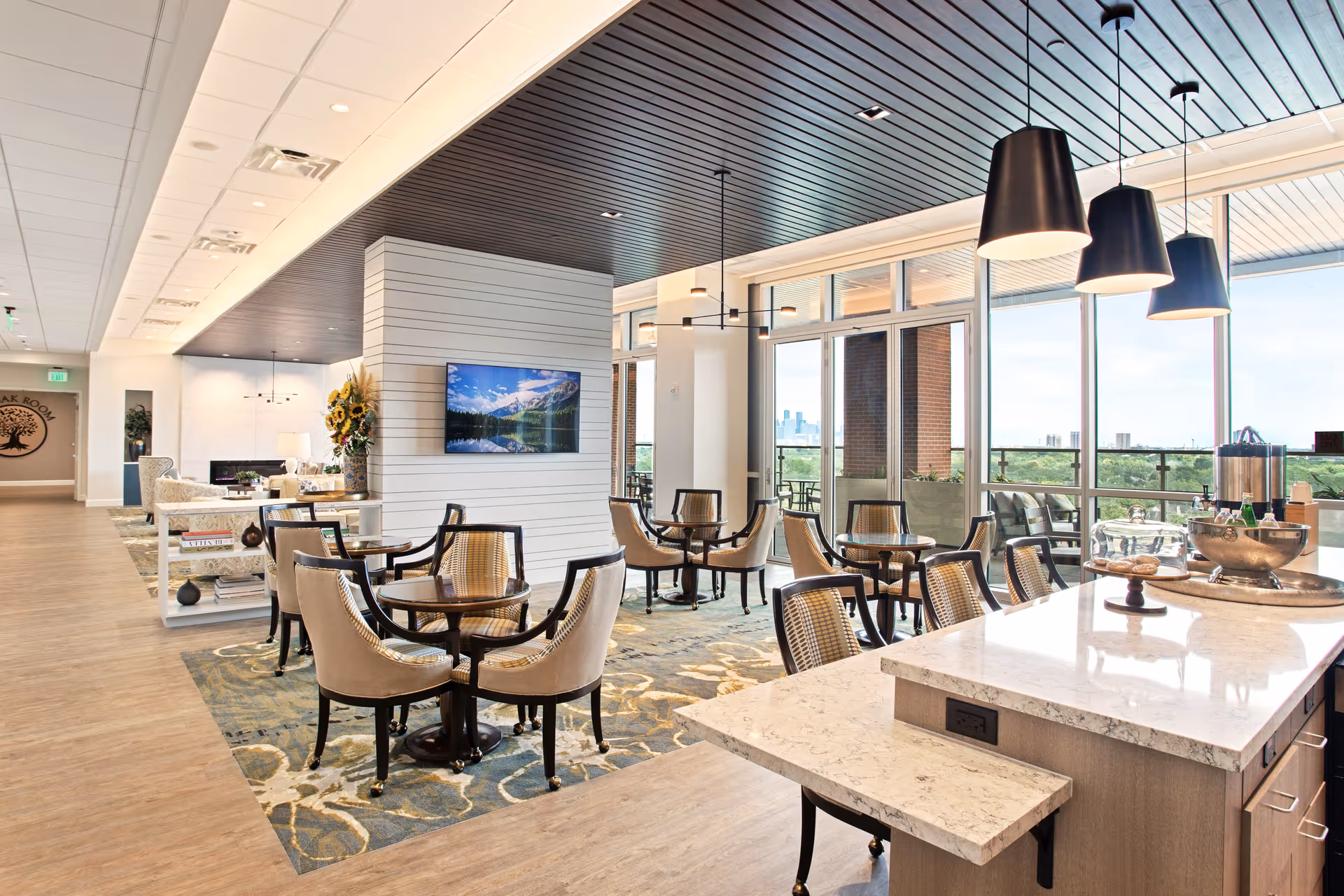Bright and modern dining area with multiple round tables and cushioned chairs on a patterned rug. A large kitchen island with marble countertop and bar stools is in the foreground. Large windows provide a view of the outdoors and allow natural light to fill the space. The ceiling has dark wood paneling with hanging pendant lights. A TV is mounted on a white shiplap wall, and there is a seating area with armchairs and a bookshelf in the background.