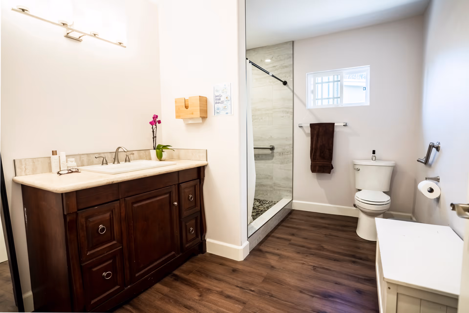 A clean and modern bathroom featuring a dark wood vanity with a beige countertop and a sink. Above the vanity is a light fixture with three lights. To the right, there is a glass-enclosed shower with a tiled interior and a small window above it. A white toilet is positioned next to the shower, with a brown towel hanging on a towel rack above it. The floor is a dark wood laminate, and there is a white storage bench on the right side of the image.