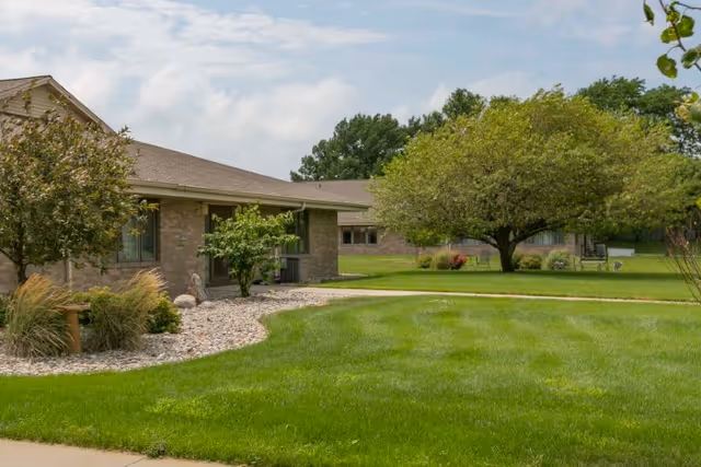 Single-story brick care facility building with a manicured lawn, trees, and a curved walkway.