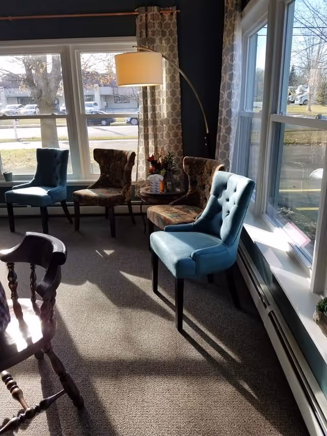 Sunlit communal sitting area with upholstered chairs, a side table and a floor lamp by large windows.