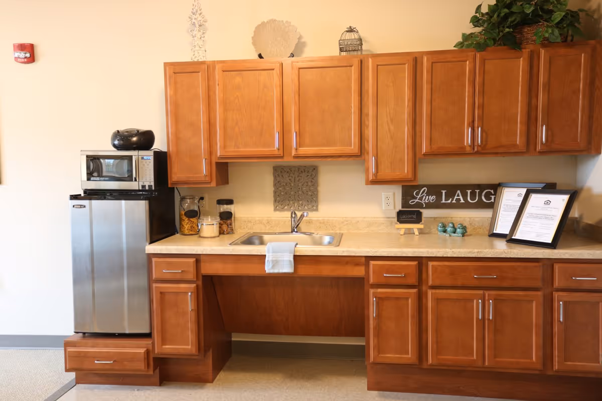 A kitchen area in Belvedere Senior Housing featuring wooden cabinets, a countertop with a sink and a towel hanging on it, a small stainless steel refrigerator with a microwave on top, jars with snacks, decorative items including a 'Live Laugh' sign, framed documents, and a potted plant on the upper cabinets.