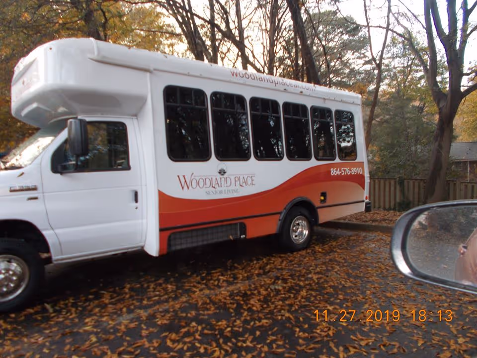 White-and-red Woodland Place senior living shuttle bus parked on a leaf-strewn street with trees and a wooden fence in the background.