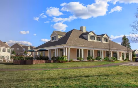 A large, light-colored building with a steep roof and dormer windows, surrounded by a well-maintained lawn and a clear blue sky with some clouds. There are small bushes planted along the building's perimeter and a paved walkway in front. Other residential buildings are visible in the background.