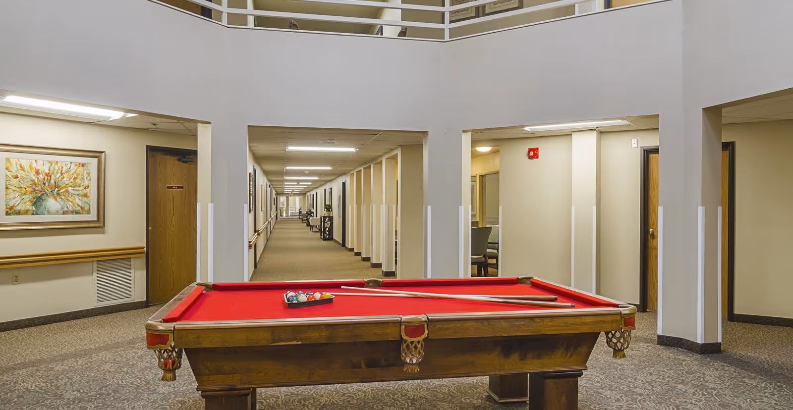Indoor common area with a red felt pool table in the foreground, surrounded by beige walls and carpeted floors. The area has a high ceiling with a balcony railing above and a long hallway extending into the background with doors and framed artwork on the walls.