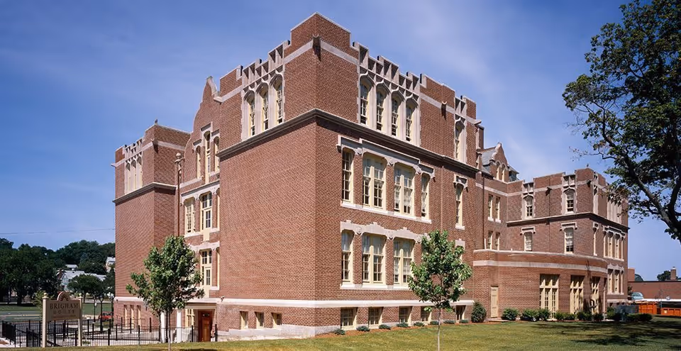 Exterior view of a large, multi-story brick building with decorative stone trim and multiple windows under a clear blue sky. There are small trees and a grassy lawn surrounding the building. A sign near the front reads 'Brigham House.'