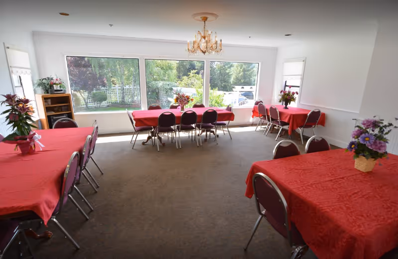 A bright dining room with large windows showing a garden outside. The room contains several tables covered with red tablecloths, each surrounded by chairs. There are flower arrangements on the tables and a chandelier hanging from the ceiling.