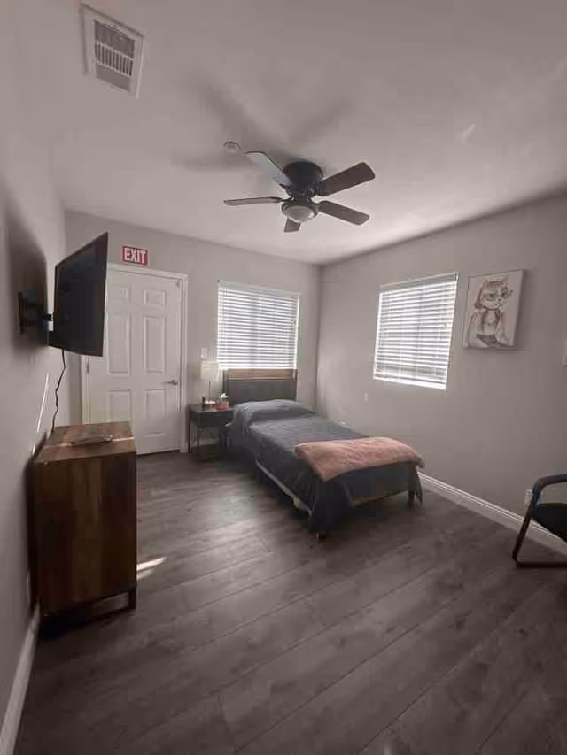A simple, clean bedroom with a single bed covered in a dark gray blanket and a pink throw at the foot. The room has two windows with blinds, a ceiling fan with light, a wall-mounted TV, a wooden cabinet, a small bedside table with a lamp, and a chair. The walls are light gray and there is an exit sign above a white door.