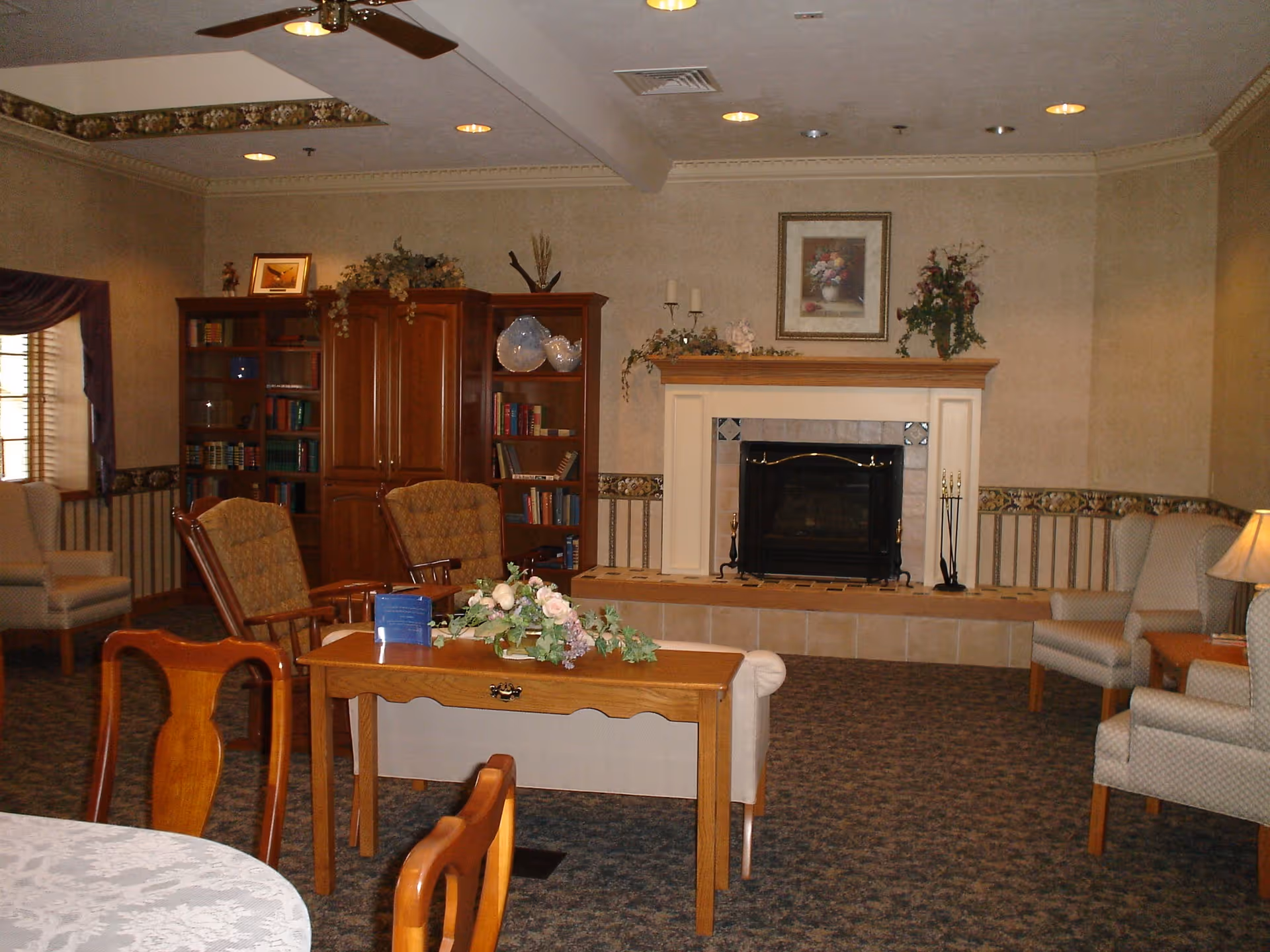 A cozy living room in a retirement community featuring a fireplace with a mantel decorated with flowers and candles. The room has several upholstered armchairs, wooden chairs, a wooden table with a floral centerpiece, a bookshelf filled with books and decorative items, and a ceiling fan. The walls are beige with a patterned border and there is a framed floral picture above the fireplace.