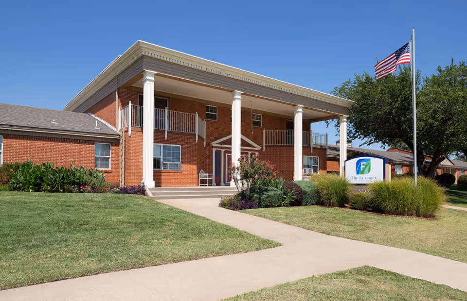 Brick two-story skilled nursing building with a covered columned entrance, manicured lawn, American flag, and a 'The Fairmont' sign.