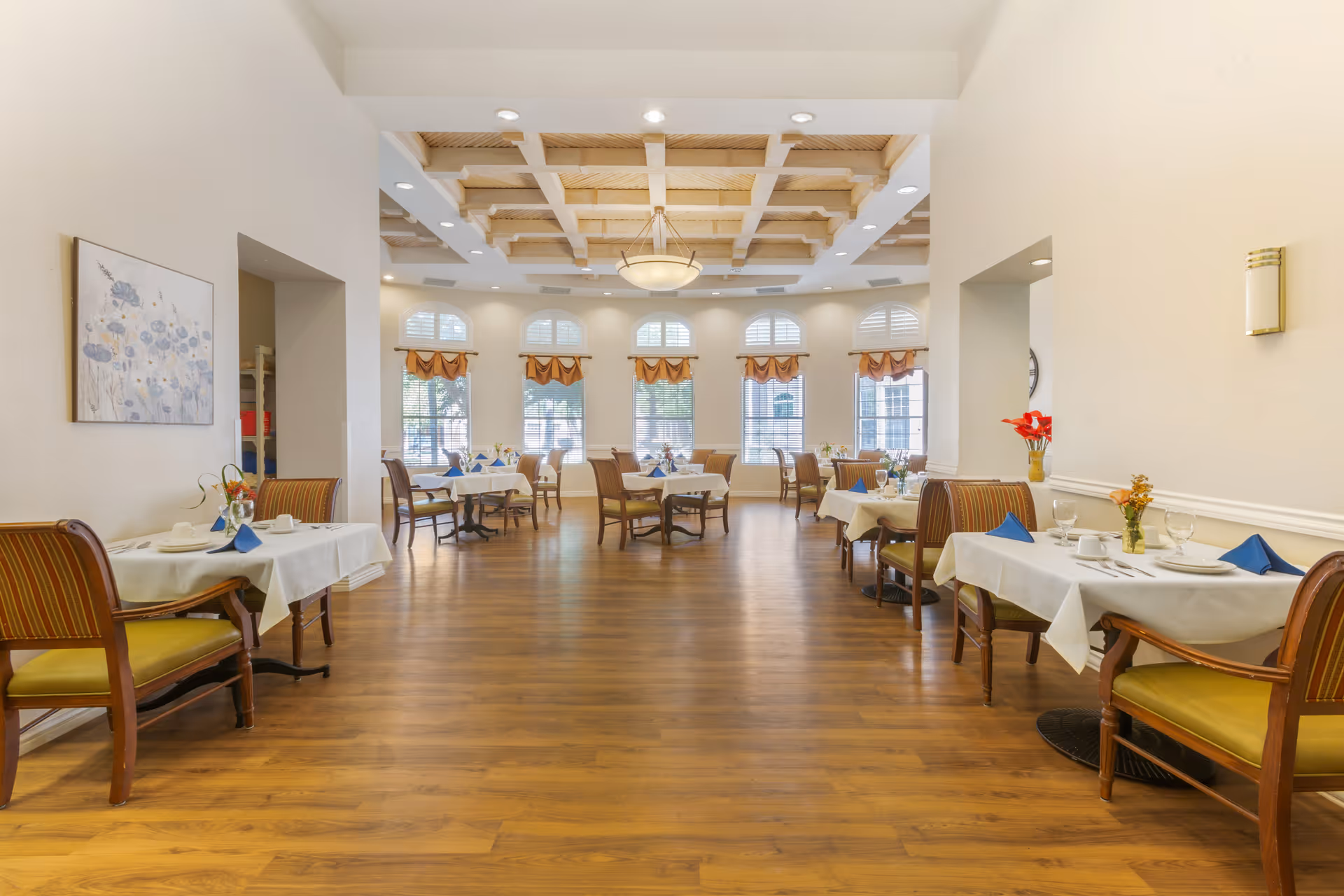 A bright and spacious dining room with wooden flooring and a decorative wooden ceiling. Several tables are set with white tablecloths, blue napkins, plates, glasses, and small flower arrangements. Large windows with white shutters and brown valances allow natural light to fill the room. The walls are painted a light cream color, and there is a painting on the left wall and a wall sconce on the right.