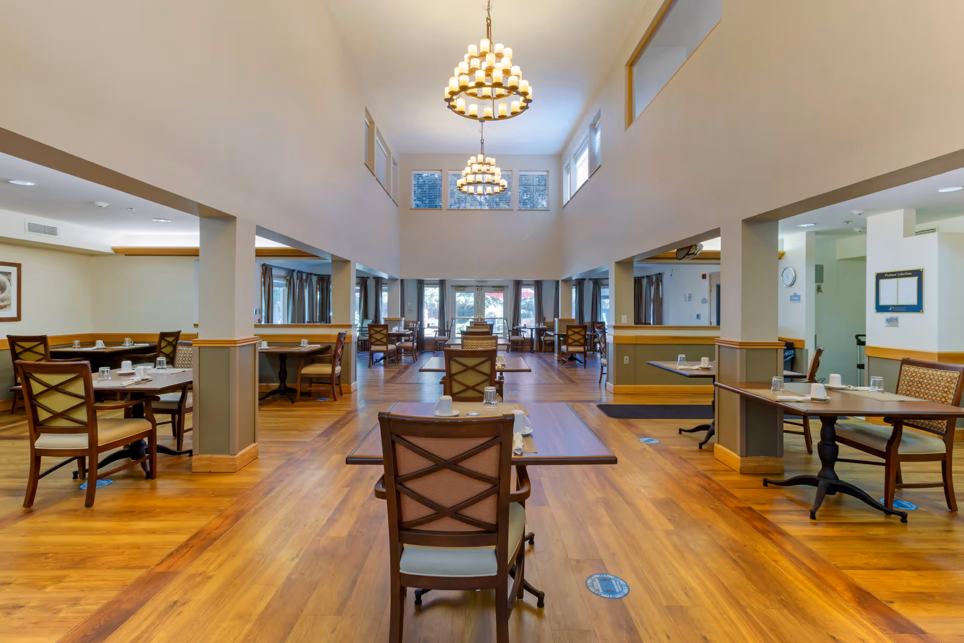 Spacious dining room with wooden floors and multiple tables set with cups and napkins. The room features high ceilings with two large chandeliers, windows near the ceiling allowing natural light, and glass doors at the far end leading outside. Chairs with cushioned seats and backs are arranged around the tables.
