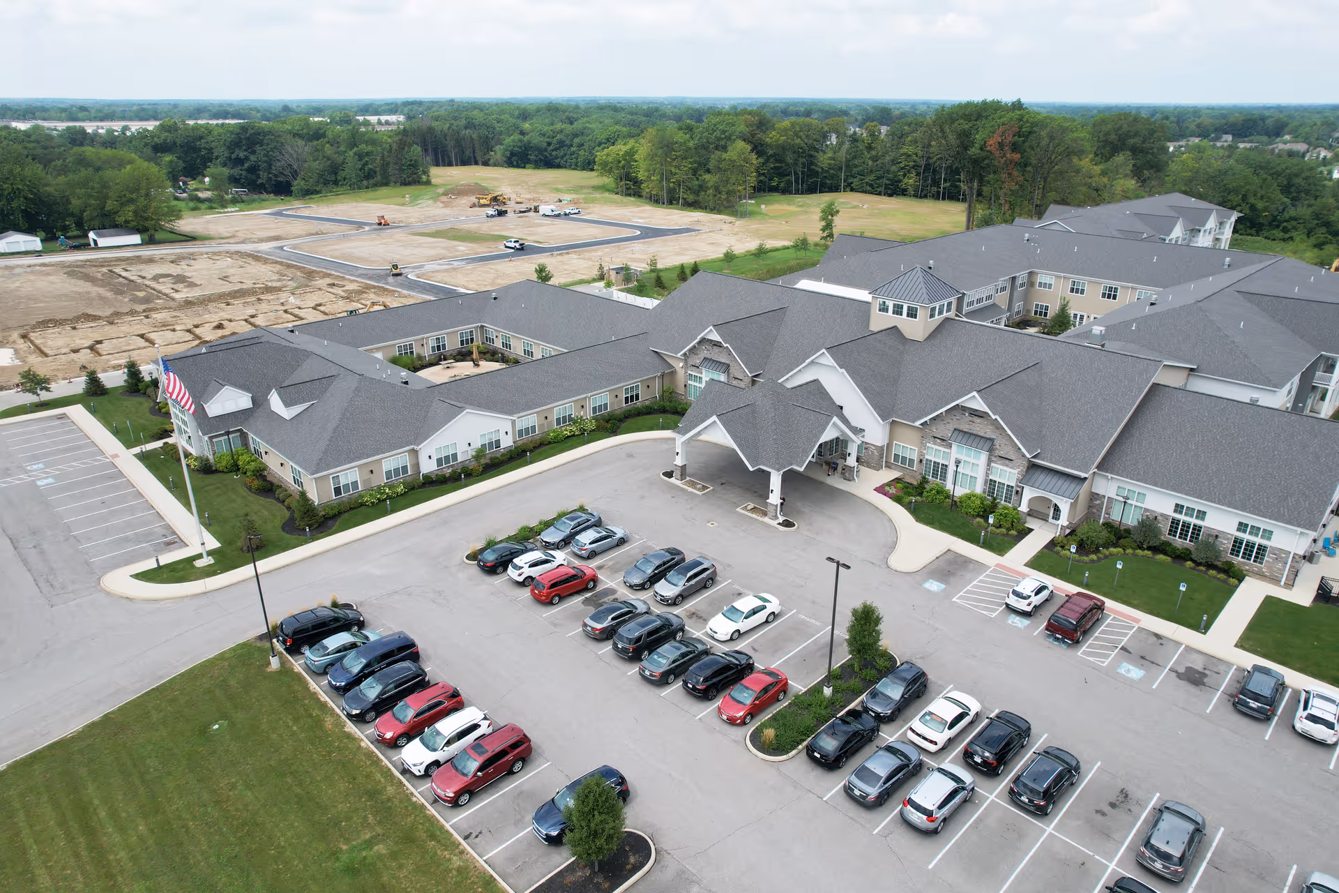 Aerial view of Vitalia Senior Residences at Strongsville showing a large senior living facility with multiple connected buildings, a covered entrance, a parking lot with numerous cars, and surrounding green areas with trees and open land.