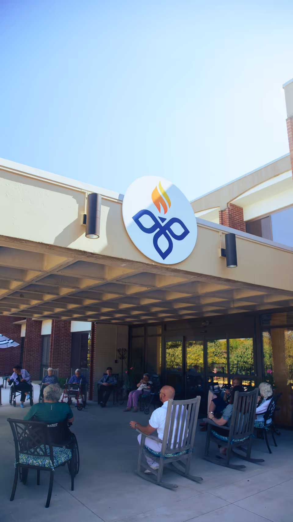 A group of elderly people sitting outside under a covered patio area of a building, some in rocking chairs and others in regular chairs, enjoying the sunny weather. The building has a large circular sign with a blue and orange flame logo above the patio.