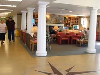 Interior view of a senior living facility common area with white columns, a stone fireplace, red armchairs, a sofa, lamps, and a few people walking and sitting in the space.