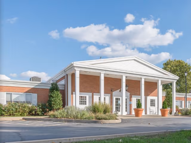 Front entrance of a single-story brick senior care facility with white columns and a portico under a blue sky.