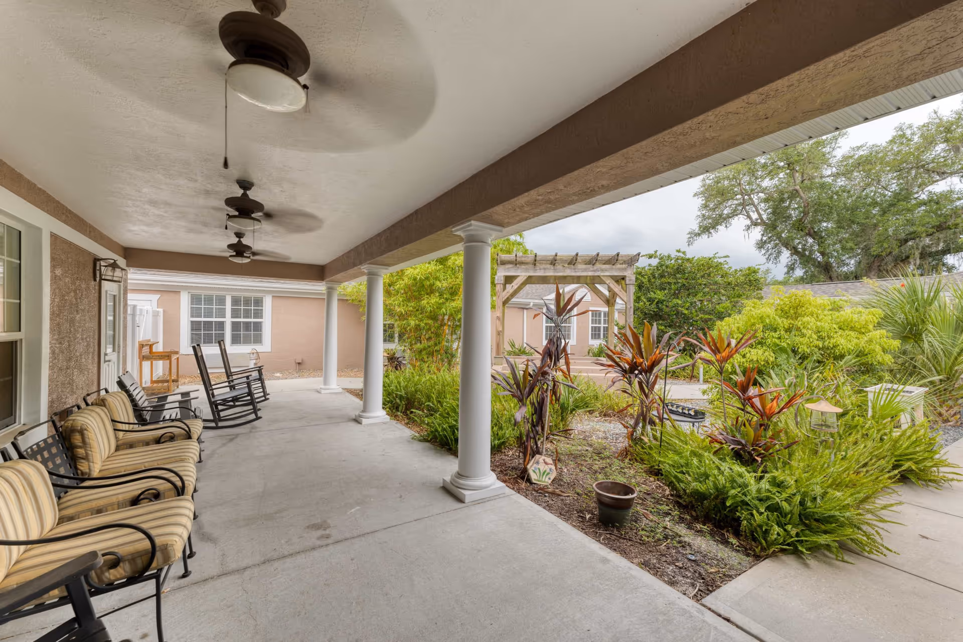 Covered outdoor patio area with cushioned chairs and rocking chairs along the wall, ceiling fans overhead, white columns supporting the roof, and a garden with various plants and a wooden pergola in the background.