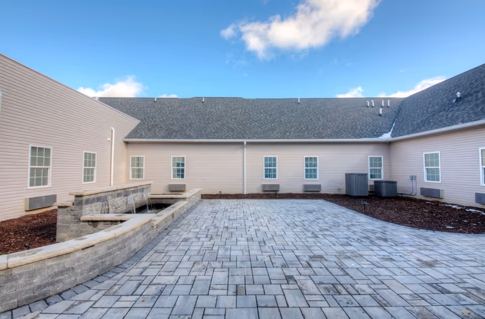 Paved courtyard with a low stone water feature in front of a beige single-story building with multiple windows and HVAC units under a blue sky.