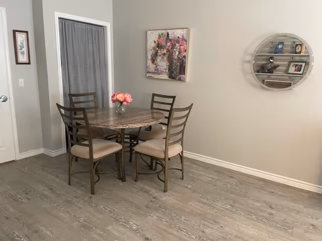 A dining area with a round table and four chairs with beige cushions. A vase with pink flowers is placed on the table. The room has light gray walls, a wooden floor, a framed floral painting on the wall, and a round wall shelf with decorative items.