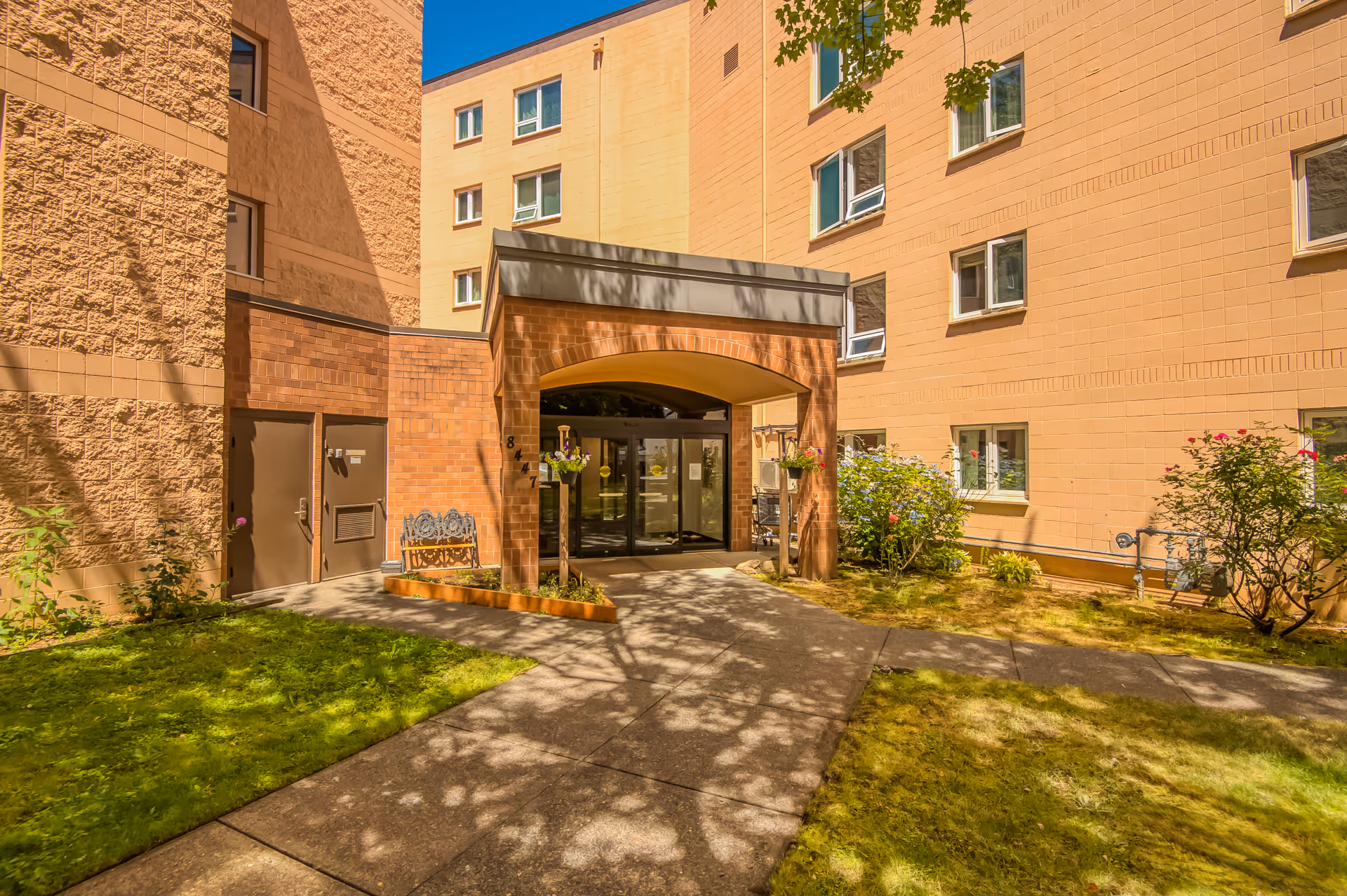Front entrance of a multi-story senior living building with a covered walkway, lawn, and shrubs.