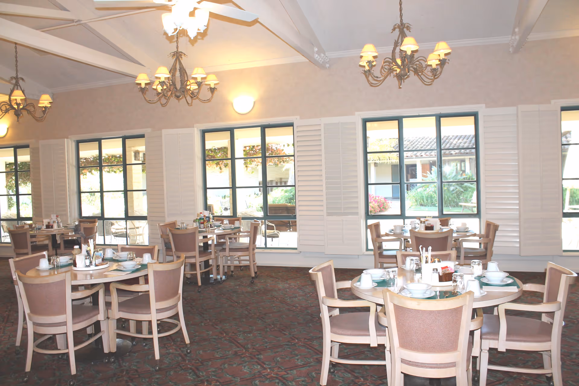 Dining room with round tables set with white dishes, cups, and condiments. The room has large windows with white shutters, chandeliers hanging from the ceiling, and a patterned carpet. Outside the windows, greenery and flowers are visible.