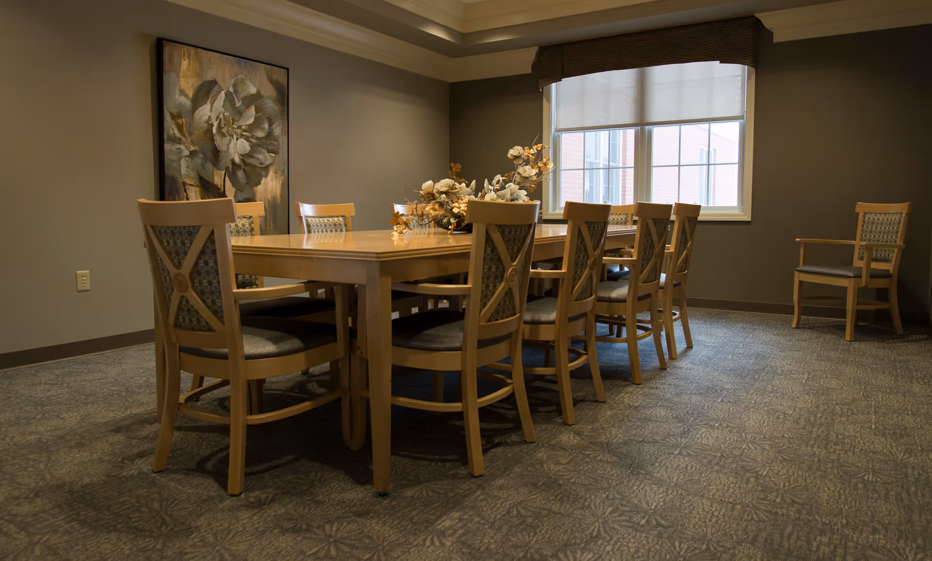 Wooden dining table surrounded by chairs with a floral centerpiece in a quiet dining room with a window and wall art.