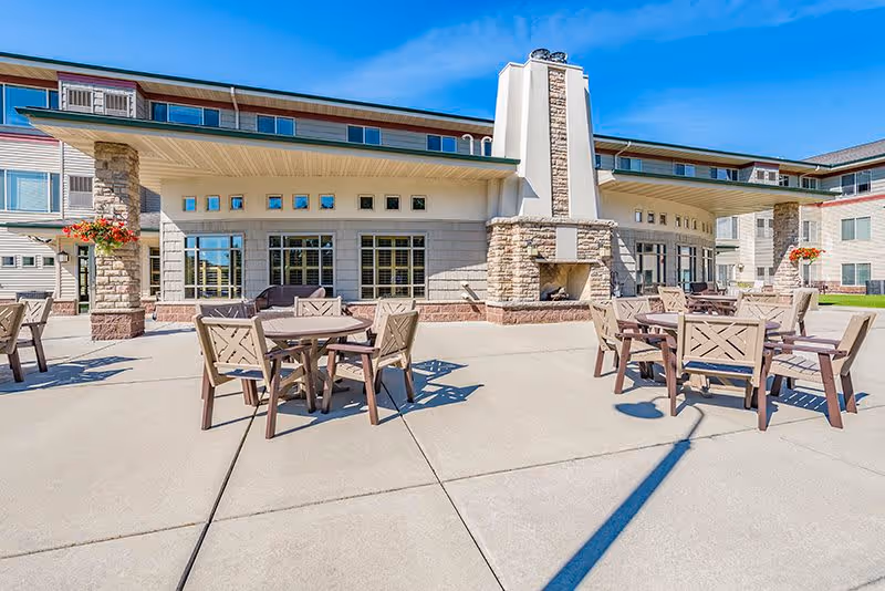 Outdoor patio area at New Perspective Senior Living | Mankato with multiple wooden tables and chairs arranged on a concrete surface. The building features large windows, stone pillars, and a central outdoor fireplace. The sky is clear and blue.