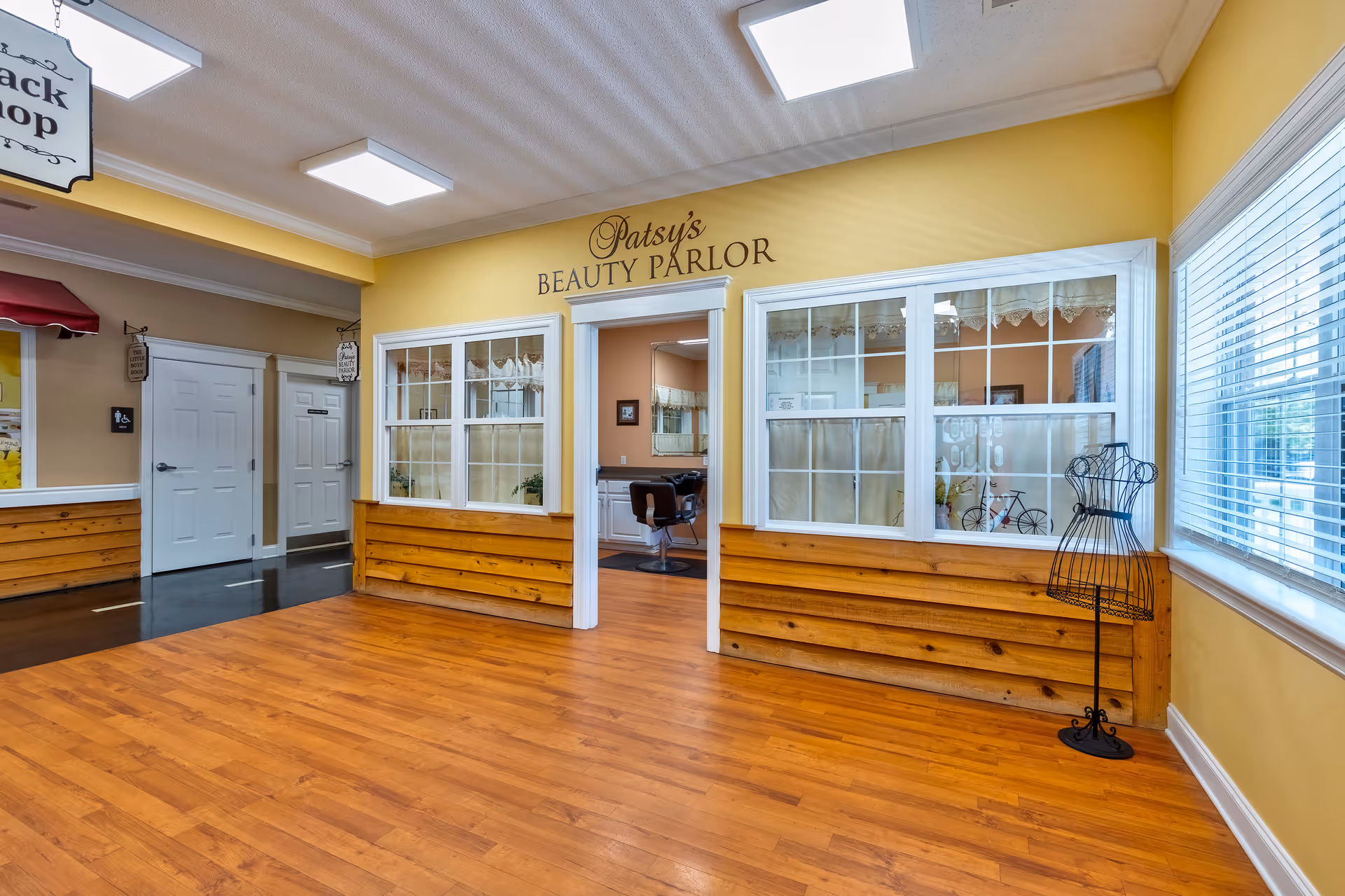 Interior hallway of a senior living facility featuring 'Patsy's Beauty Parlor' storefront with large windows, wooden wainscoting, and laminate flooring.