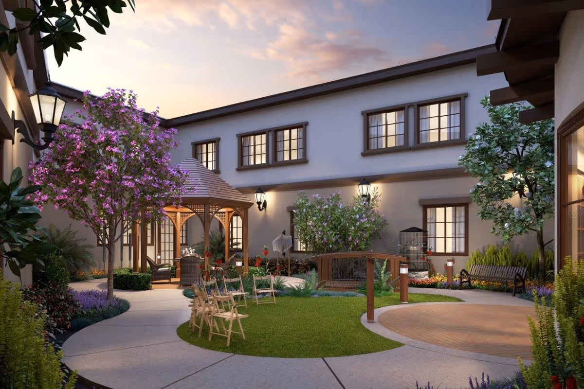 A peaceful outdoor courtyard at sunset with a wooden gazebo, several wooden chairs arranged on a circular grassy area, a small wooden bridge over a garden bed, flowering trees, shrubs, and a bench along the pathway. The courtyard is surrounded by a two-story building with multiple windows and wall-mounted lanterns providing warm light.