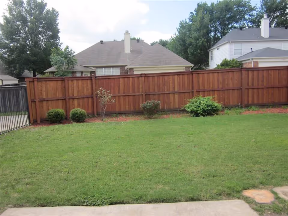 Grassy backyard with a wooden privacy fence, small shrubs, and neighboring houses visible beyond.