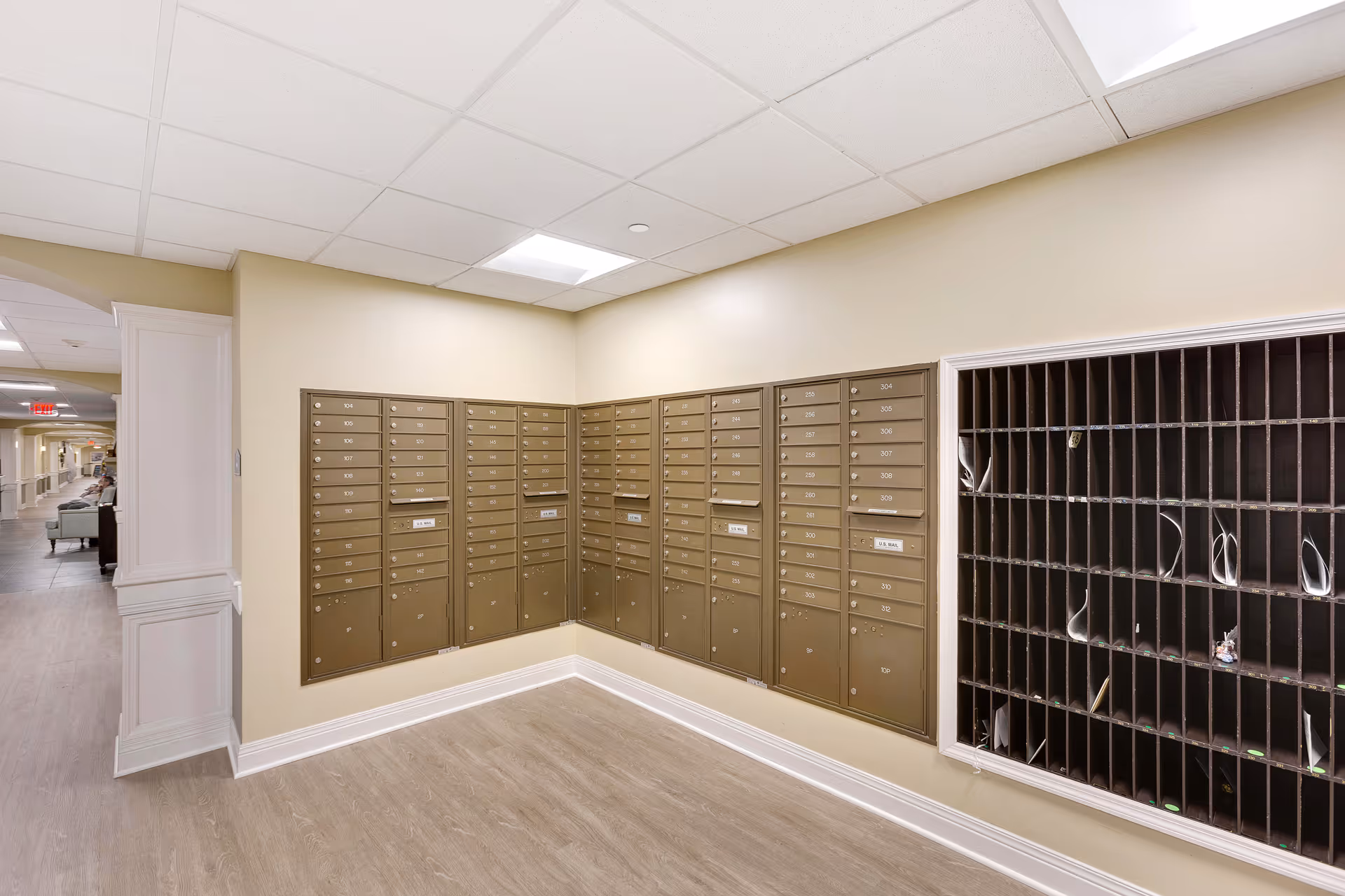 Interior view of a senior living facility mailroom with multiple rows of beige mailboxes mounted on the walls and a large sorting cubby unit on the right side. The floor has light wood-style flooring, and the walls are painted a light beige color. A hallway with seating is visible in the background to the left.