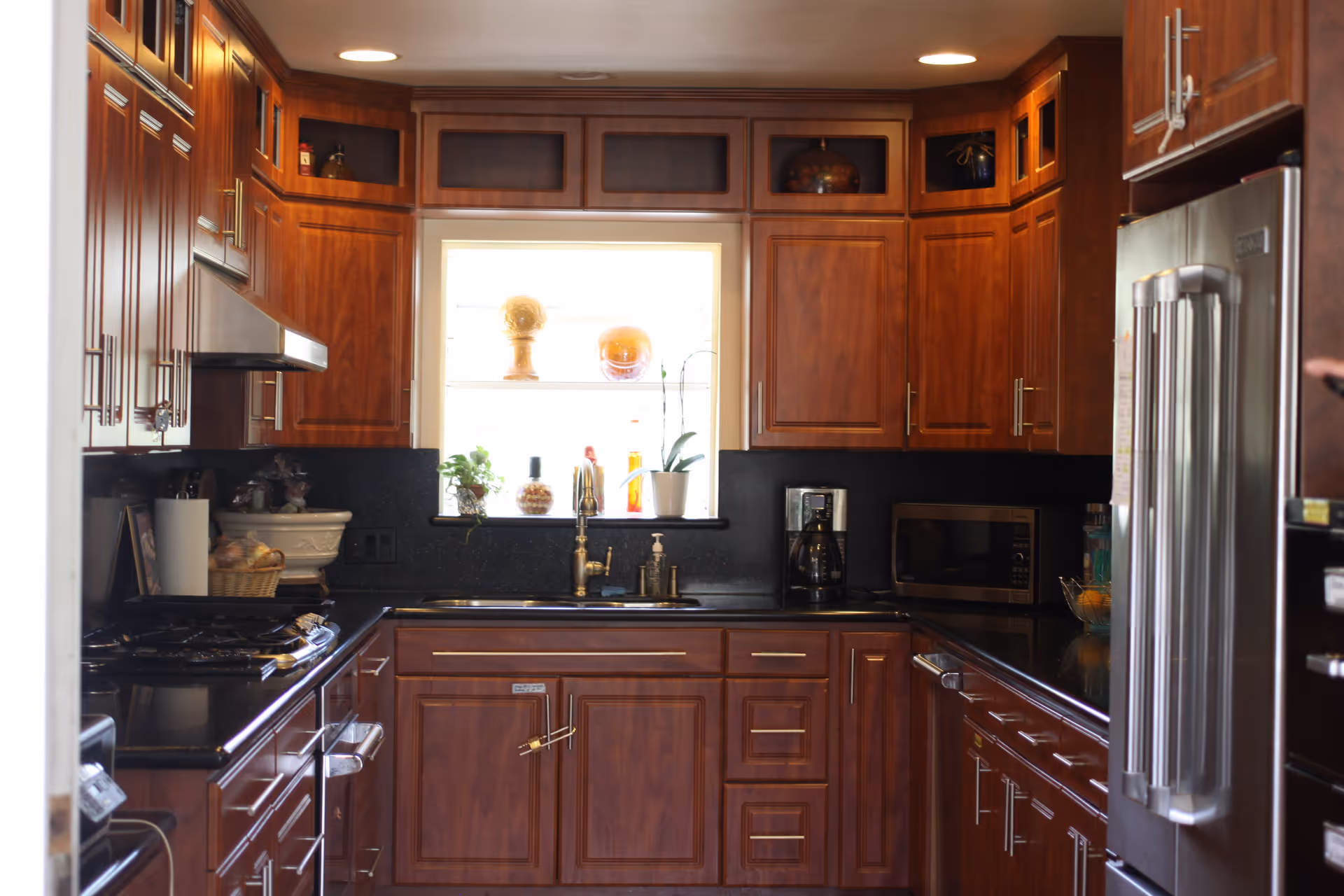 A kitchen with dark wooden cabinets, black countertops, a stainless steel refrigerator, a microwave, a coffee maker, and a window above the sink with decorative items on the windowsill.