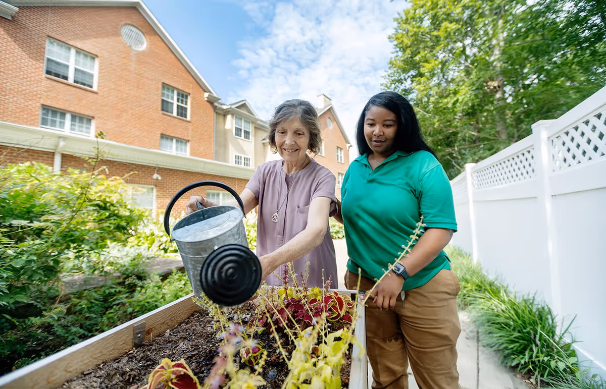 An elderly woman watering plants in a raised garden bed outdoors with the assistance of a younger caregiver. They are in a garden area next to a brick building and a white fence, under a partly cloudy sky.