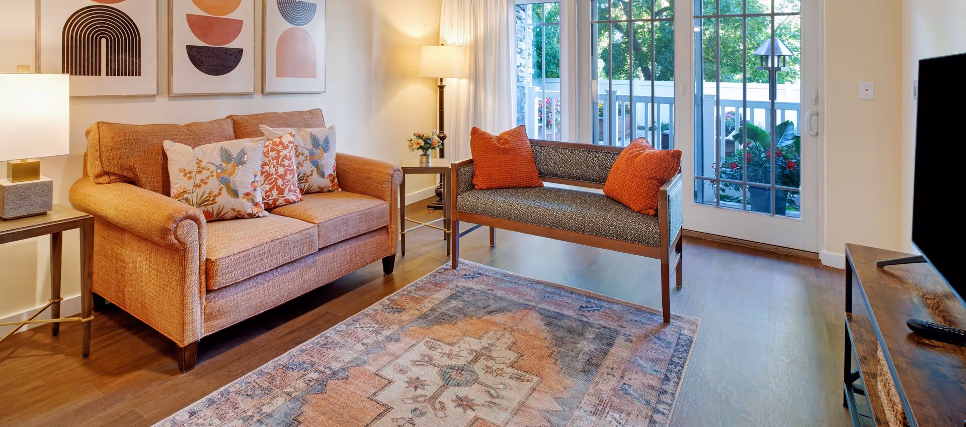 Bright living room with an orange loveseat, patterned rug, bench with orange pillows, and glass doors opening to a balcony.