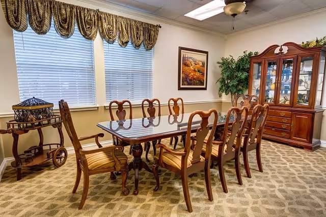 A formal dining room with a long wooden table and eight matching chairs with striped cushions. The room features a patterned carpet, two large windows with blinds and decorative valances, a wooden china cabinet filled with dishes, a framed landscape painting on the wall, a potted plant in the corner, and a wooden serving cart.