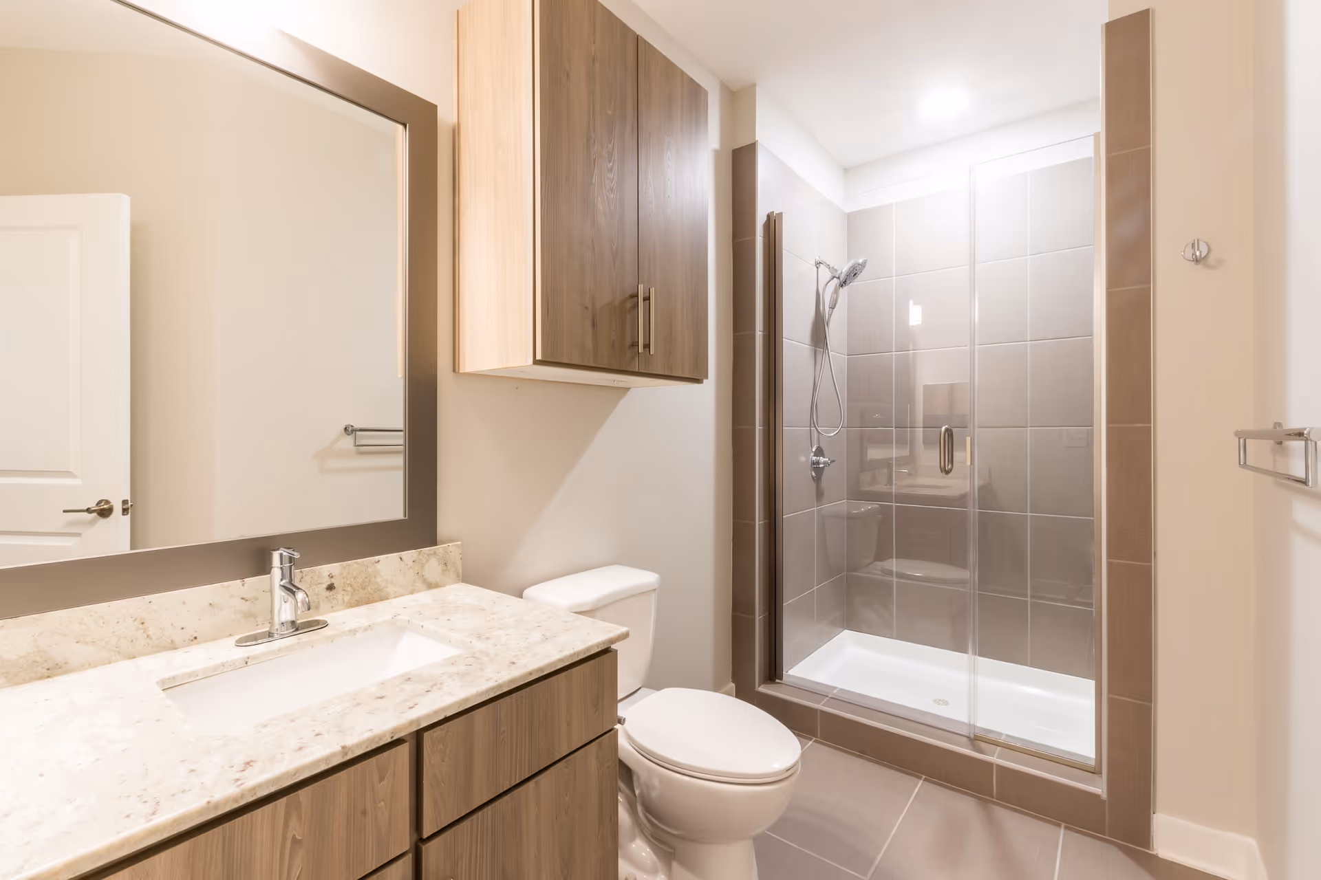A modern bathroom featuring a large mirror above a granite countertop with a sink, wooden cabinets below and above the toilet, and a glass-enclosed shower with gray tiles and a handheld showerhead.