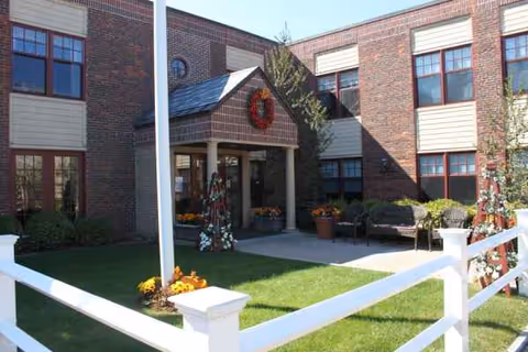 Brick senior living facility entrance with wreath, benches, manicured lawn, and white fence.