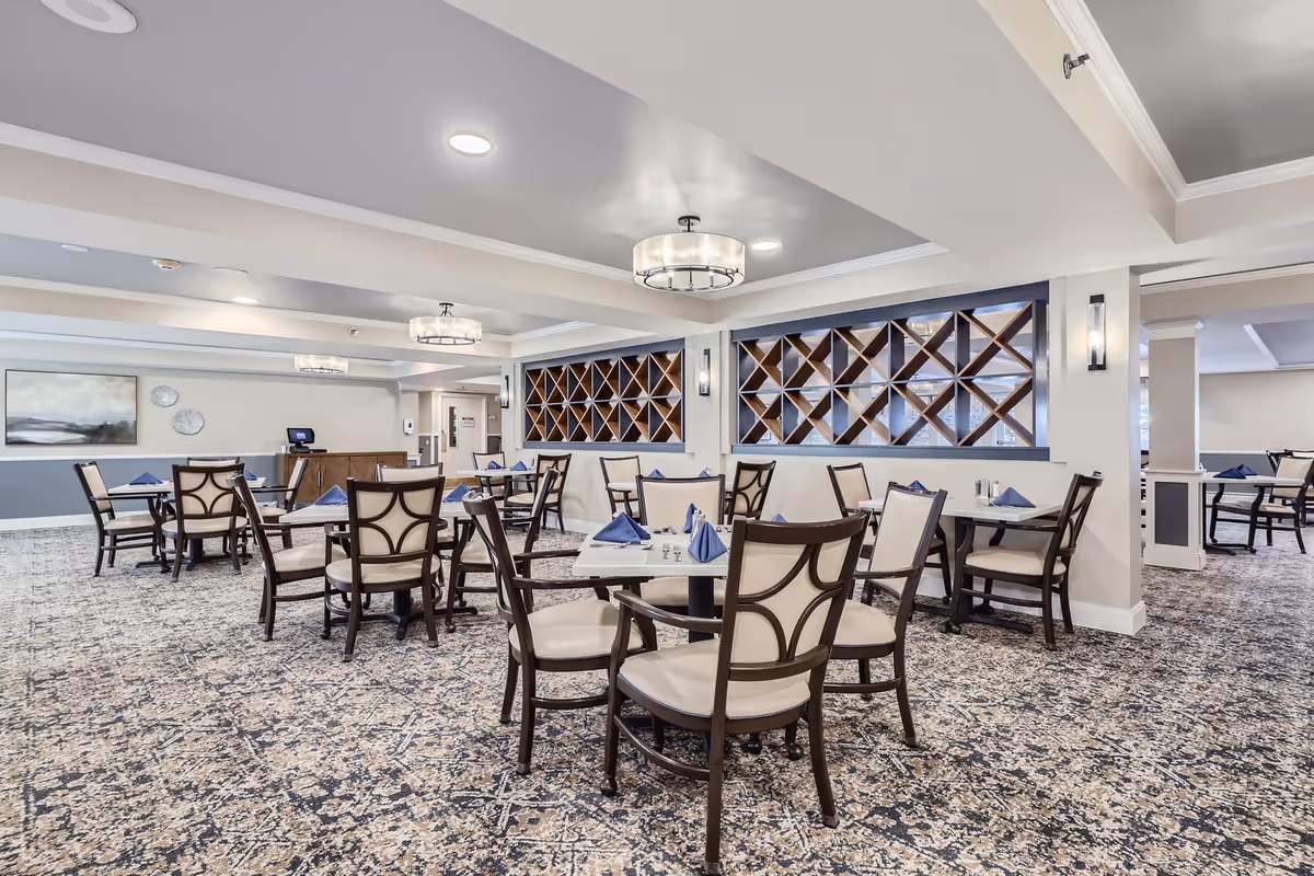A spacious dining room with multiple tables and chairs arranged neatly. Each table is set with blue folded napkins and silverware. The room features patterned carpet flooring, modern ceiling lights, and decorative wooden shelving on the walls. The walls are painted in light neutral tones with some artwork and clocks visible in the background.