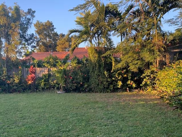 A grassy lawn bordered by tropical palm trees, shrubs, and flowering plants with a red-tiled roof visible behind a fence.