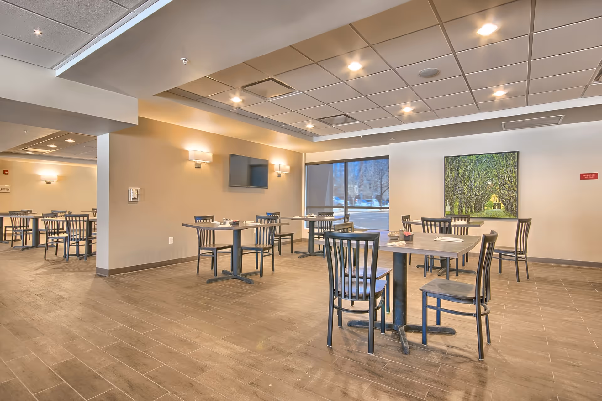 A spacious dining room with several tables and chairs arranged neatly. The room has a tiled floor, beige walls, and a drop ceiling with recessed lighting. There is a large window letting in natural light, a wall-mounted TV, and a painting of a tree-lined path on one wall.