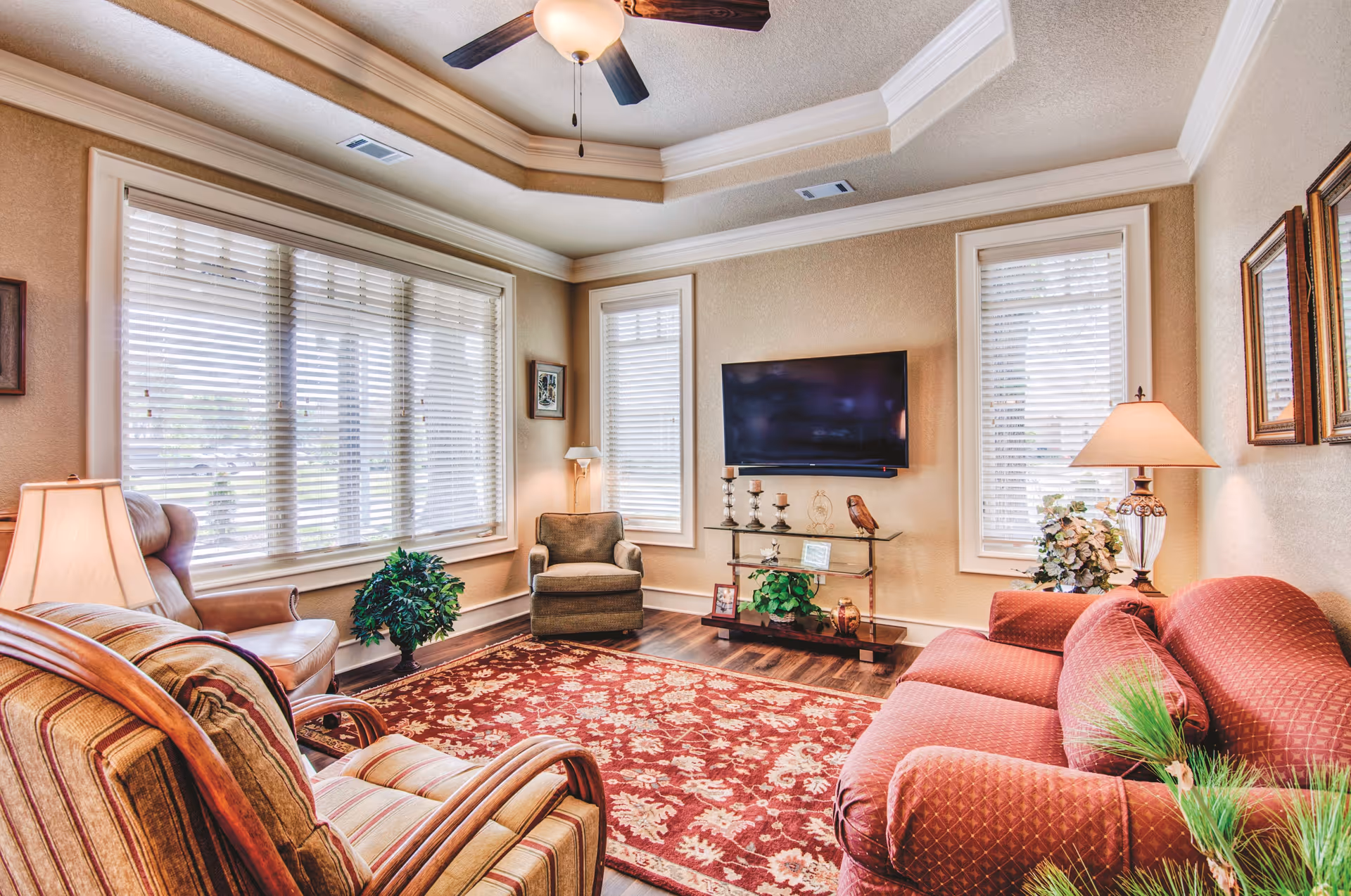 A cozy living room with a red patterned sofa, two armchairs, a large window with blinds, a ceiling fan, a flat-screen TV mounted on the wall, decorative plants, lamps, and a red floral area rug on wooden flooring.