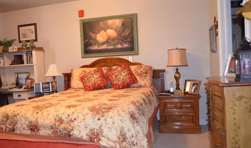 A cozy bedroom in a senior living facility featuring a large wooden bed with floral bedding and two red decorative pillows. On the wall above the bed is a framed painting of white roses. To the left of the bed is a white bookshelf with various decorative items and a lamp on a small table. To the right of the bed is a wooden nightstand with a lamp, framed photo, and other small items. A wooden dresser with a decorative lantern and picture frame is visible on the right side of the image.