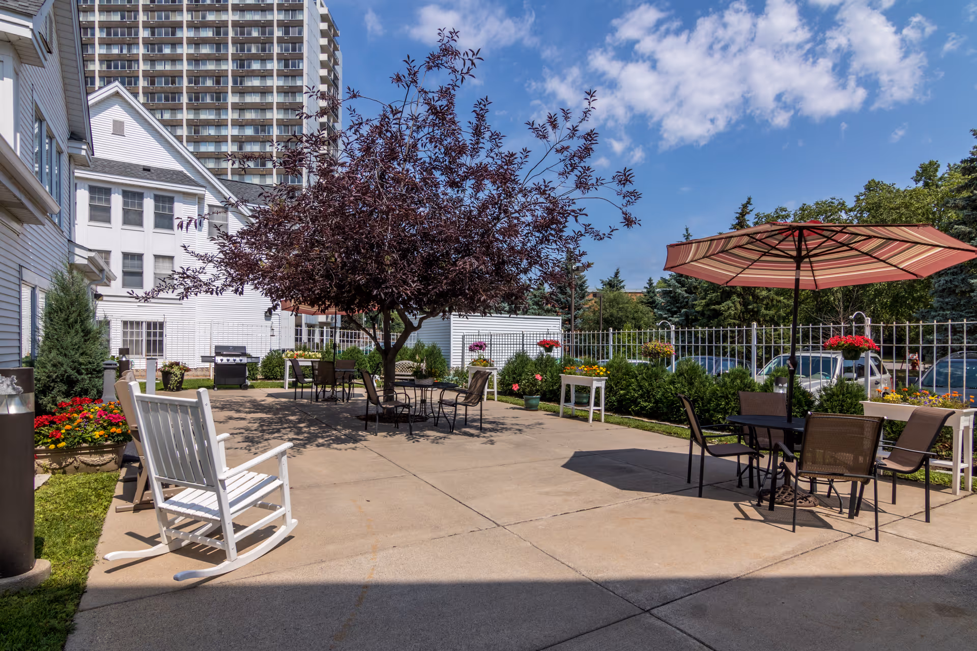 Outdoor courtyard patio with tables, chairs, a striped umbrella, a white rocking chair, potted flowers, and a central tree next to a senior living building.
