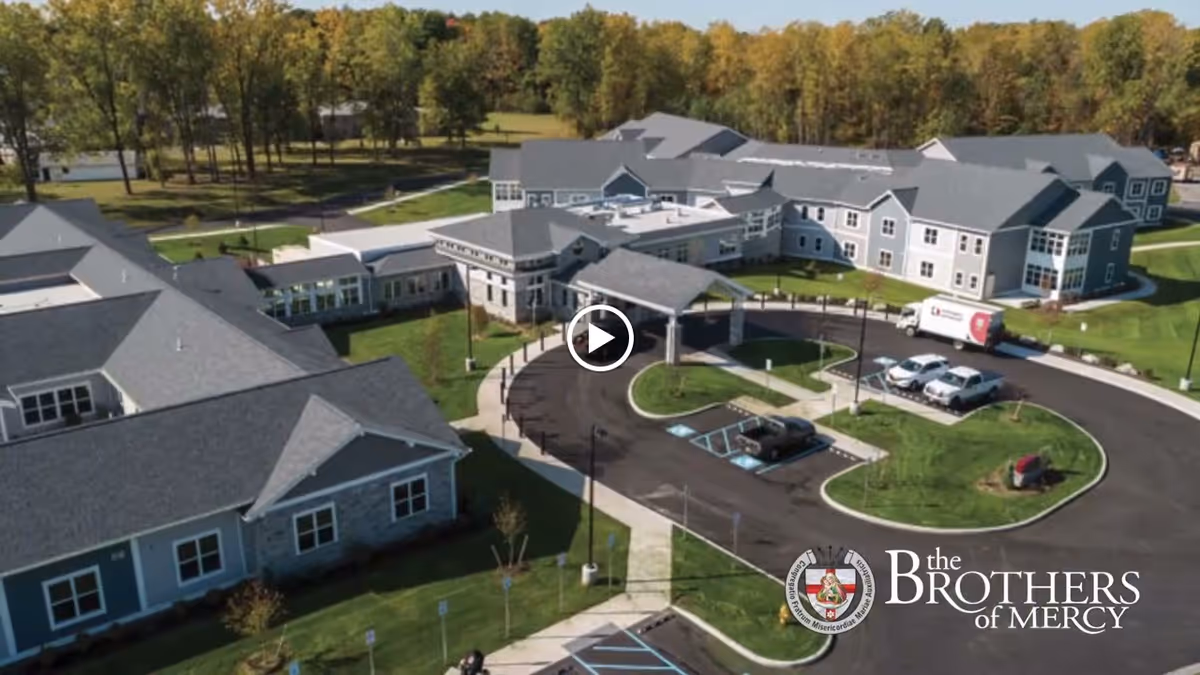 Aerial view of the Brothers of Mercy senior living facility showing multiple connected buildings with gray roofs, a circular driveway with parked vehicles, green lawns, and trees in the background under a clear sky.