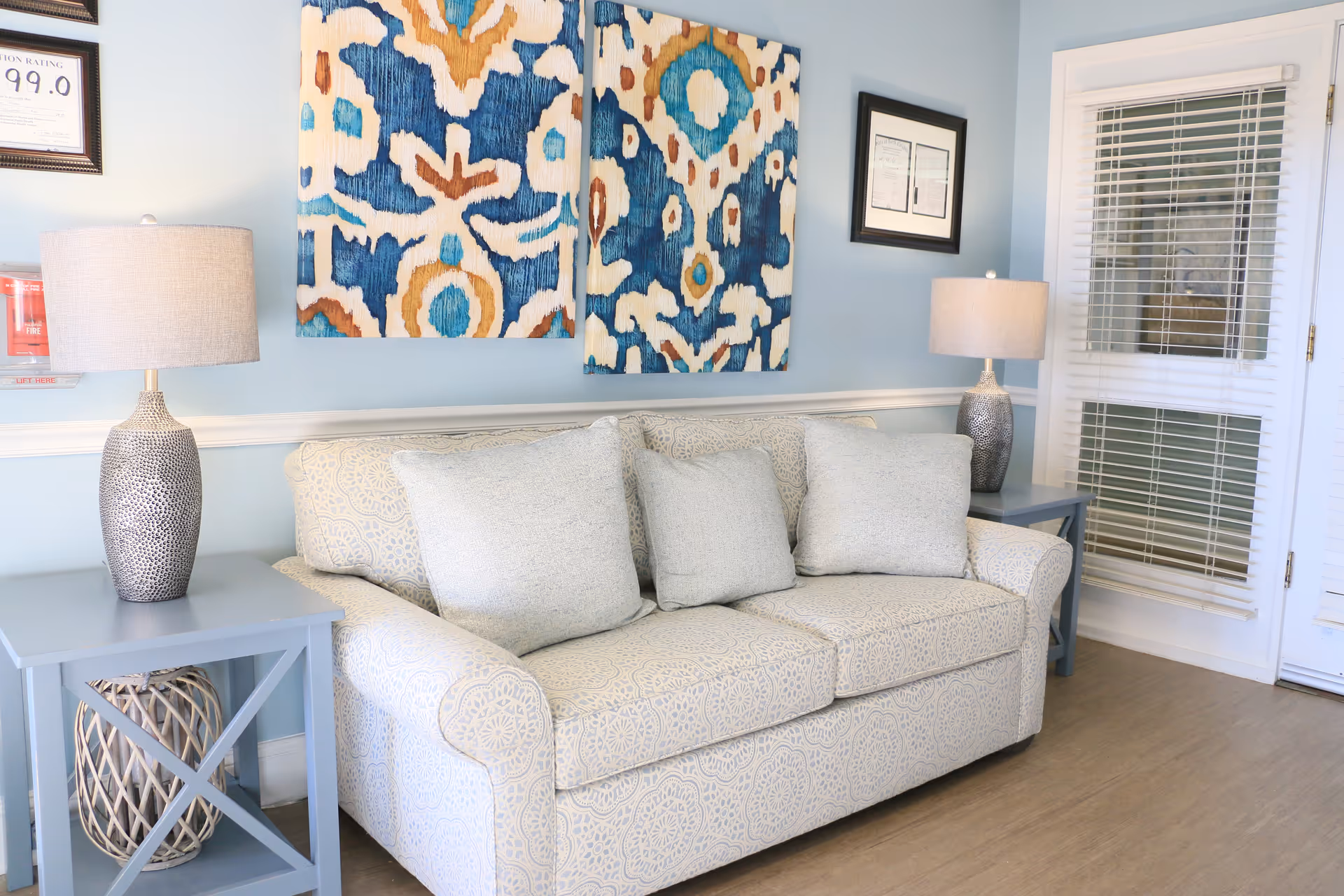Light-blue living room with a patterned sofa, two side tables with lamps, and colorful abstract wall art.