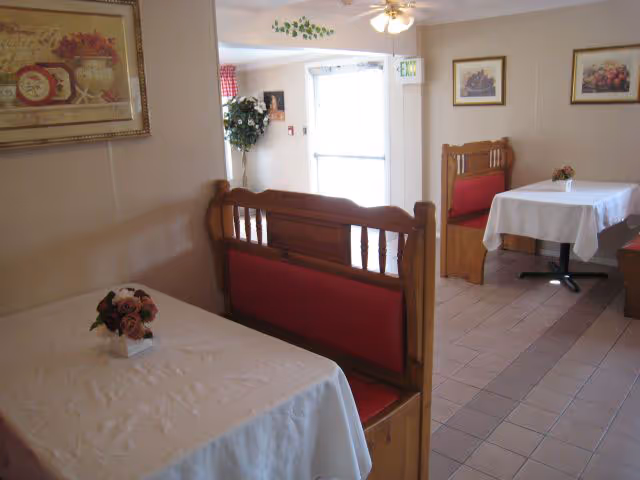 Small dining area with wooden booths and tables covered in white tablecloths, floral centerpieces, framed artwork on the walls, and an exit door.