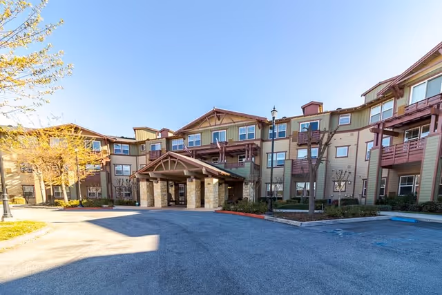 Exterior view of a three-story senior living facility building with a covered entrance supported by stone pillars, multiple windows, balconies, and a clear blue sky above. The surrounding area includes a paved driveway, some trees, and landscaping.