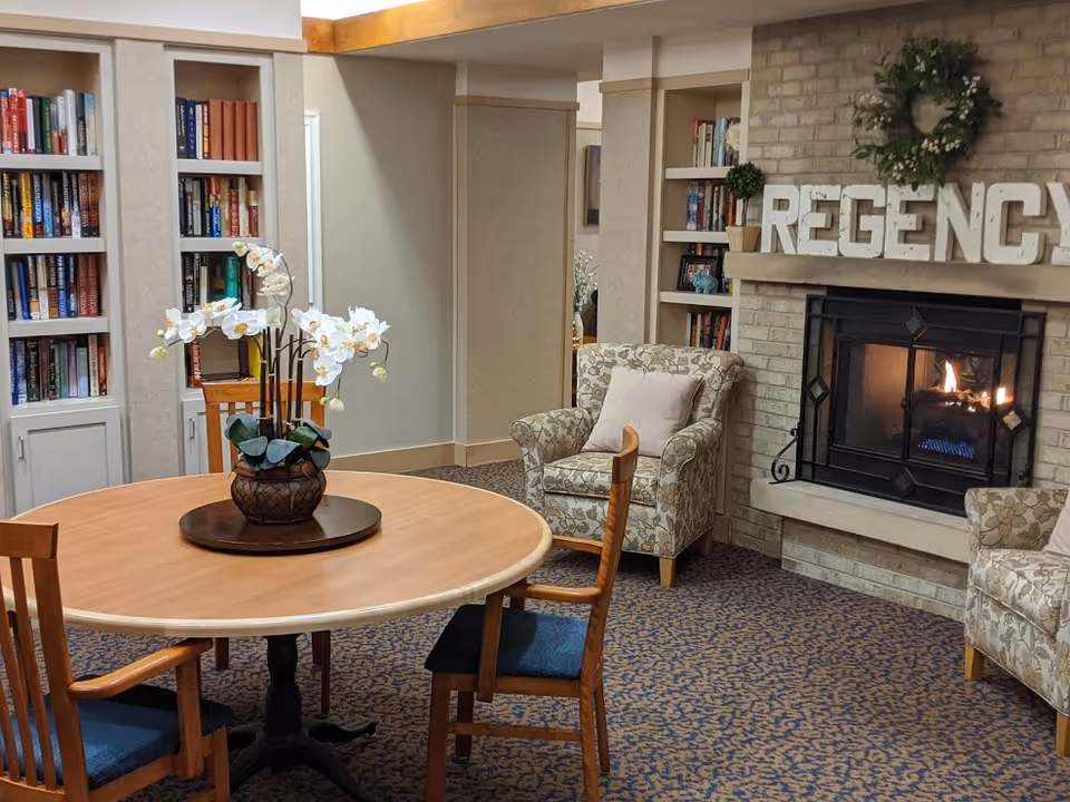 A cozy senior living common room with a round table and chairs, a floral centerpiece, bookshelves, armchairs, and a lit fireplace with 'REGENCY' letters above it.